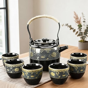 Op de afbeelding: Een zwarte keramische theeset bestaande uit een theepot met een gebogen handvat omwikkeld met lichtgekleurde vezels en vijf bijpassende kopjes. Elk stuk heeft een goudkleurig bloemmotief en een gestippelde textuurband aan de bovenrand.