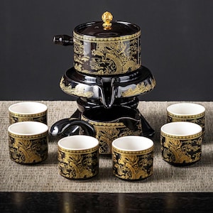 Könnte beinhalten: Schwarzes und goldenes Keramik-Teeservice mit Drachenmotiv. Das Set umfasst eine Teekanne, eine Servierkanne und sechs kleine Tassen. Die Teekanne hat einen Deckel mit einem goldenen Knopf. Die Tassen und Kannen haben ein goldenes Drachendesign.