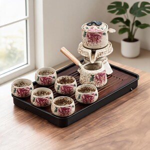 May include: A ceramic tea set with a floral design. The set includes a teapot, a serving pot with a wooden handle, and six small cups. The tea set is placed on a dark wooden tray. The floral design features pink peonies.