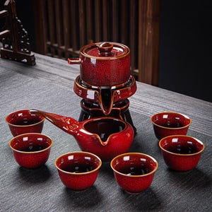 May include: A red ceramic tea set with a teapot, serving pot, and six small cups. The set has a crackled glaze and gold trim. The teapot sits on a stand, and the serving pot has a long spout.
