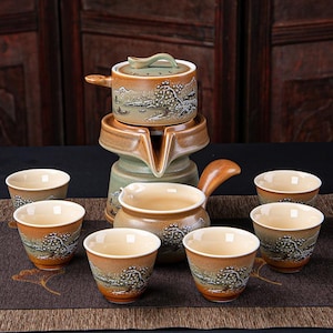 May include: A ceramic tea set with a landscape design. The set includes a teapot, a server, and six teacups. The teapot and cups have a brown and beige colour scheme with a painted tree and mountain scene.