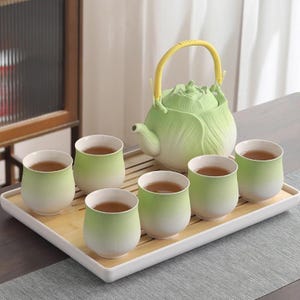 May include: A green ceramic tea set with a teapot and six cups, all filled with tea. The teapot has a yellow handle and a cabbage-like design. The cups are arranged on a bamboo tray.
