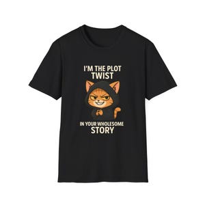 May include: Black t-shirt featuring a cartoon cat wearing a hooded cloak. The cat has orange and brown fur and a mischievous expression. Text on the shirt reads "I'M THE PLOT TWIST IN YOUR WHOLESOME STORY."
