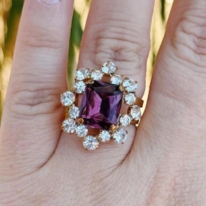 May include: A gold-toned ring featuring a large, square-cut, deep purple central stone. The central stone is surrounded by a halo of twelve small, round, clear white stones set in a floral-like arrangement on a metallic band.