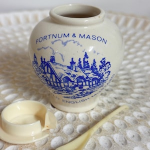 May include: A cream-coloured ceramic jar with a blue printed illustration of a cottage and the text FORTNUM & MASON and OLD ENGLISH MUSTARD. The jar includes a matching cream-coloured plastic lid and a small cream-coloured plastic spoon.