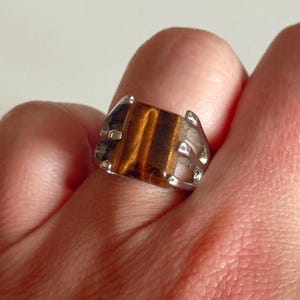 May include: A silver ring with a rectangular tiger's eye stone. The stone displays brown and gold bands. The ring has a decorative silver setting with openwork details on the sides. The ring is shown on a finger.