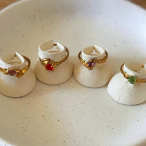 May include: Four gold-toned rings, each featuring a single small round gemstone in pink, red, purple, and green. The rings are displayed on individual white rounded stands, showcasing a simple band design with a prong setting for each stone.