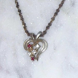 May include: A silver-toned metal necklace featuring a heart-shaped pendant with a swirling design. The pendant is accented with two small round red stones and three clear white stones arranged in a diagonal line across the centre of the heart.