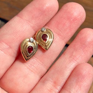 Pode incluir: Um par de brincos de ouro, cada um com um design em forma de lágrima, uma pedra preciosa vermelha central em corte de pera e uma pequena pedra de destaque redonda e clara no topo. Os brincos têm um acabamento polido e uma estrutura metálica em camadas.