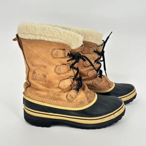 May include: Pair of tan leather Sorel Caribou winter boots with black rubber soles and cream-colored faux fur trim. The boots have black laces and metal eyelets. The Sorel logo is visible on the side.