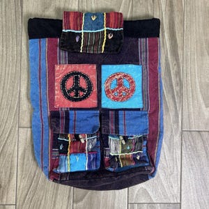 May include: A colorful, patchwork backpack with a peace sign design. The bag features a mix of blue, red, and purple striped fabric, with multiple pockets and a flap closure. The peace signs are in red and blue squares.