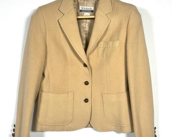 Könnte beinhalten: Ein beigefarbener Blazer mit klassischem Design, mit fallendem Revers, zwei Vordertaschen und drei Knöpfen an jeder Manschette. Die Jacke hat einen Dreiknopfverschluss und zwei Vordertaschen. Der Blazer hängt an einem Holzkleiderbügel.