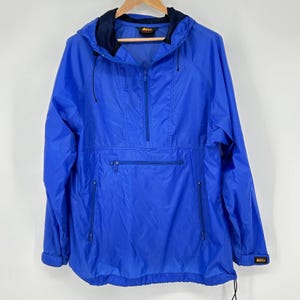 May include: A bright blue anorak-style jacket with a hood and a half-zip closure. The jacket has a front pocket, two side pockets, and the REI logo. The jacket is hanging on a wooden hanger against a white background.