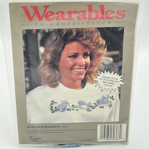 Op de afbeelding: Een kruissteekpakket genaamd "Wearables" met een vrouw die een witte sweatshirt draagt met geborduurde blauwe vogels en bloesems. De kit bevat 2,54 cm tear-away canvas en katoenen floss. De tekst "Design Done on 2,54 cm Tear Away Canvas" is ook zichtbaar.
