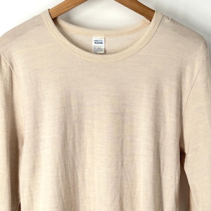 Könnte beinhalten: Langärmeliges T-Shirt mit Rundhalsausschnitt in hellem Beige. Das T-Shirt hängt an einem Holzkleiderbügel. Der Stoff scheint ein weiches, leichtes Material zu sein. Ein Etikett ist in der Nähe des Halsausschnitts sichtbar.