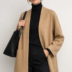 Puede incluir: Una mujer con un cárdigan color camel sin cierre, un cuello alto negro y pantalones negros. Lleva un bolso tote negro. El conjunto es sencillo y elegante, adecuado para un entorno profesional o informal.