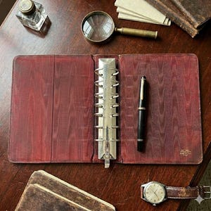 May include: An open, burgundy leather binder with a silver ring mechanism, a black pen, and a vintage wristwatch with a brown leather band. A magnifying glass, inkwell, and small stack of paper are also visible on a wooden surface.