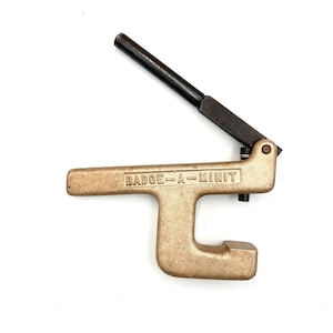 May include: A metallic gold-toned Badge-A-Minit manual press tool featuring a textured finish and a long, black cylindrical handle attached to the top. The brand name Badge-A-Minit is embossed in capital letters on the side of the main frame.