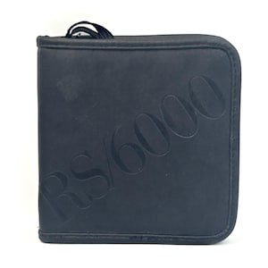 May include: A square black zippered case featuring the embossed text RS/6000 across the front surface. The case has rounded corners, a matte finish, and a small black fabric loop attached to the top edge for carrying or hanging purposes.