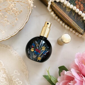 May include: A round, dark-colored glass perfume bottle with a gold-toned spray nozzle and a matching gold cap. The bottle features a painted floral design with blue, red, and yellow flowers and green leaves. The bottle is positioned upright.