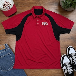 Könnte beinhalten: Ein rotes Poloshirt mit schwarzen Schulterpartien und einem Teamlogo auf der Brust. Eine passende Baseballkappe und schwarz-weiße Turnschuhe sind ebenfalls zu sehen. Das Shirt hat einen Kragen und einen Knopfverschluss.