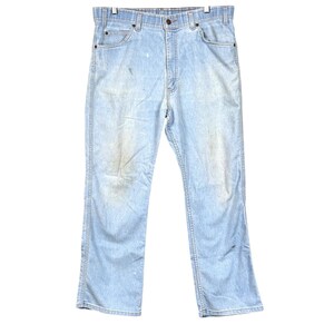 May include: Light blue denim jeans featuring a classic five-pocket design, a button fly closure, and belt loops. The fabric displays a faded wash with subtle distressed markings and light wear patterns across the front of the legs.