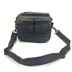 Puede incluir: Una bolsa para cámara de color negro con un cuerpo rectangular estructurado, un asa superior y una correa de hombro de nailon negro ajustable y extraíble. La solapa frontal presenta una costura central vertical y un pequeño logotipo.