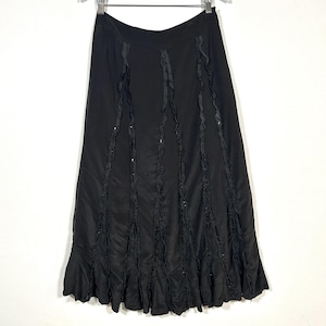 May include: Black, floor-length skirt with vertical ruffled accents and a flared hem. The skirt is hanging on a hanger against a white background. The ruffles have a subtle sheen, adding texture to the garment.