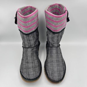 May include: A pair of gray and black winter boots. The boots have a textured pattern and a gray cuff with pink stripes. The cuffs are folded over and secured with a black strap. The soles are brown.