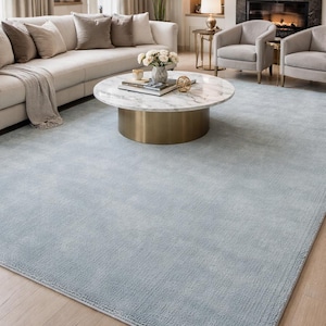 May include: A light blue area rug in a living room setting. The rug is rectangular and covers most of the floor. A white sofa with beige pillows, a marble-topped coffee table, and two armchairs are visible.