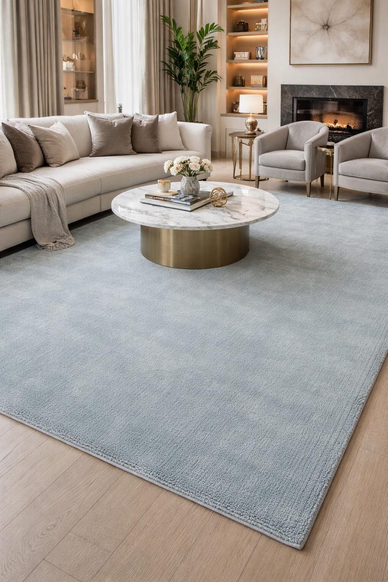 May include: A light blue area rug in a living room setting. The rug is rectangular and covers most of the floor. A white sofa with beige pillows, a marble-topped coffee table, and two armchairs are visible.