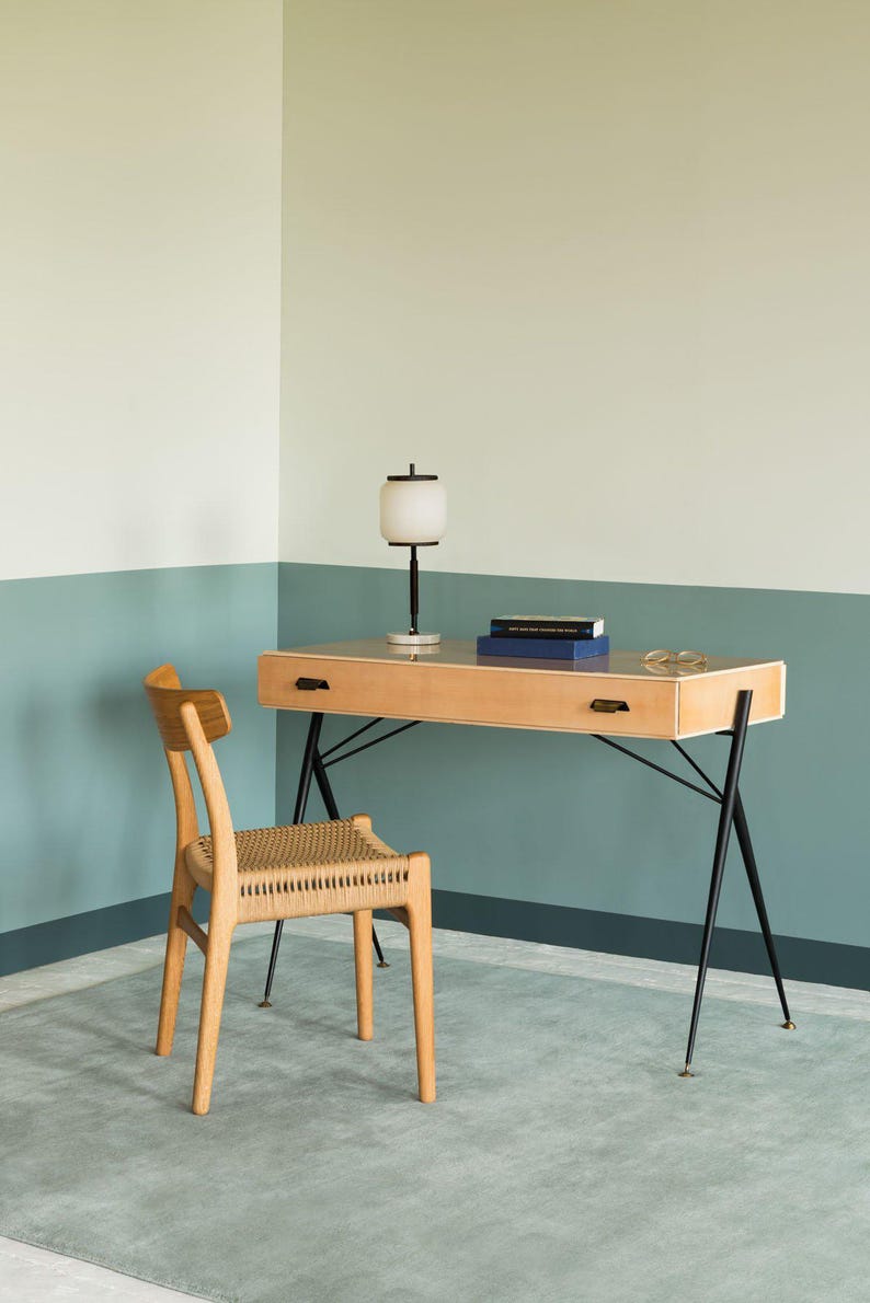 May include: A light wood desk with black metal legs and a matching chair with a woven seat. A white and black lamp sits on the desk, along with two books and a pair of glasses. The room has a two-tone wall in light green and blue.