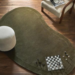Può includere: Un tappeto verde oliva di forma organica con un bordo più scuro. Una piccola scacchiera quadrata con pezzi degli scacchi è posizionata sul tappeto. Un pouf bianco e una sedia con una rivista sono anche nell'immagine.