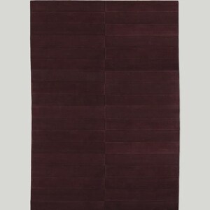 May include: A rectangular, burgundy-colored rug with a geometric design. The rug is divided into rectangular sections, creating a textured, striped appearance. The rug is a solid color, with no patterns or additional details.