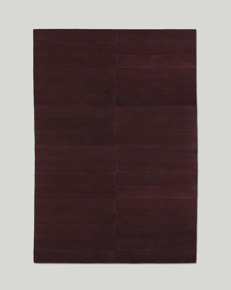 May include: A rectangular, burgundy-colored rug with a geometric design. The rug is divided into rectangular sections, creating a textured, striped appearance. The rug is a solid color, with no patterns or additional details.