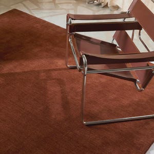 Può includere: Scena d'interni moderna con un tappeto color ruggine e una sedia in pelle marrone con struttura cromata. Il tappeto copre una vasta area del pavimento e la sedia è posizionata sopra. L'estetica generale è minimalista e contemporanea.