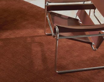 May include: A modern interior scene featuring a rust-colored rug and a brown leather chair with a chrome frame. The rug covers a large area of the floor, and the chair is positioned on top of it. The overall aesthetic is minimalist and contemporary.