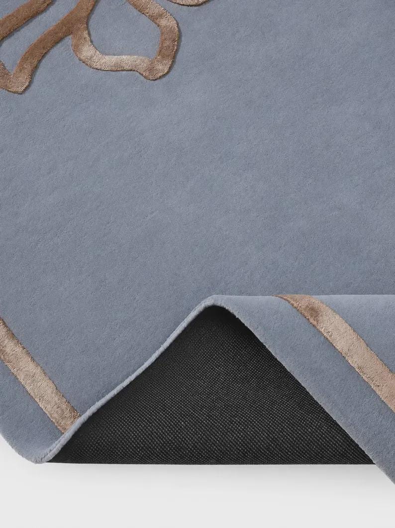 Pu&ograve; includere: Un tappeto blu con un motivo floreale in rilievo color bronzo metallizzato e bordi lineari. Il tappeto &egrave; parzialmente ripiegato per rivelare un materiale di supporto nero, testurizzato e antiscivolo sul lato inferiore della superficie.