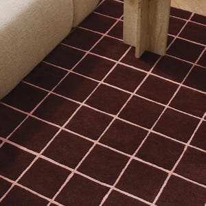 May include: A dark brown area rug with a grid pattern of light pink lines. The rug is rectangular and placed on a wooden floor. A beige sofa and a small, round side table are partially visible in the background.