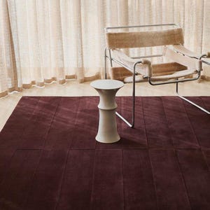 Può includere: Un tappeto rettangolare bordeaux con una superficie strutturata. Uno sgabello decorativo bianco è sul tappeto. Una sedia moderna con struttura in metallo e tessuto beige è sullo sfondo. Tende color crema sono dietro la sedia.