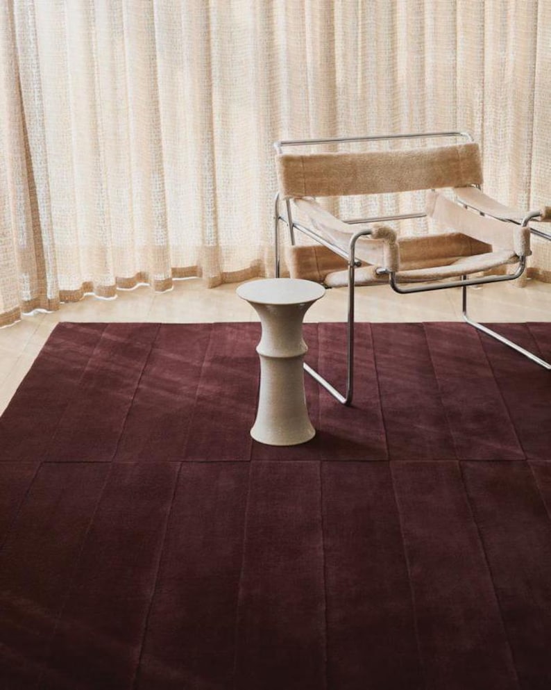 May include: A burgundy rectangular rug with a textured surface. A white, decorative stool stands on the rug. A modern chair with a metal frame and tan fabric is in the background. Sheer, cream-colored curtains are behind the chair.