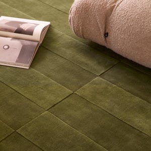 Peut inclure: Gros plan sur un tapis vert avec un motif géométrique. Le tapis est vert olive foncé et a une surface texturée. Un fauteuil beige texturé est partiellement visible en arrière-plan, ainsi qu'un magazine ouvert.