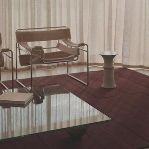 May include: A modern living room scene featuring a burgundy rug, a glass-topped coffee table, and a beige chair with a metal frame. A small, white, hourglass-shaped side table stands nearby, with beige curtains in the background.