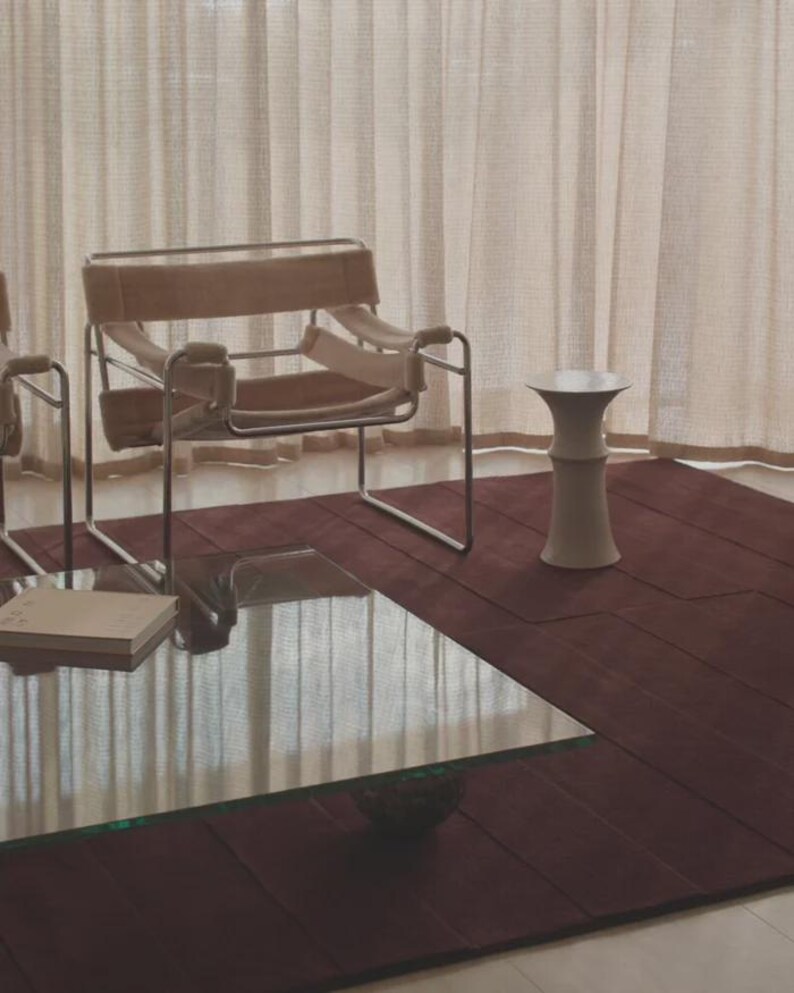May include: A modern living room scene featuring a burgundy rug, a glass-topped coffee table, and a beige chair with a metal frame. A small, white, hourglass-shaped side table stands nearby, with beige curtains in the background.