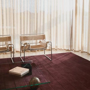May include: A modern living room scene featuring a deep burgundy rug with a geometric pattern. Two chairs with metal frames and tan upholstery are visible. A glass coffee table holds a book and a decorative sphere. Beige curtains provide a soft backdrop.