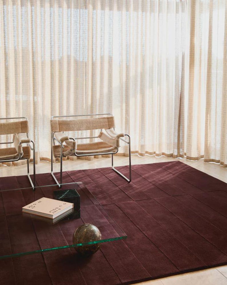 May include: A modern living room scene featuring a deep burgundy rug with a geometric pattern. Two chairs with metal frames and tan upholstery are visible. A glass coffee table holds a book and a decorative sphere. Beige curtains provide a soft backdrop.