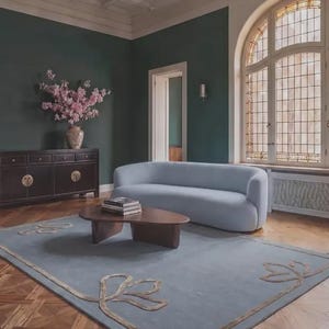Pu&ograve; includere: Un divano curvo azzurro poggia su un grande tappeto blu con un bordo floreale color marrone chiaro. Un tavolino da caff&egrave; in legno marrone scuro con una pila di libri &egrave; posto davanti al divano, vicino a un mobile scuro con fiori rosa.