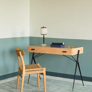 May include: A light wood desk with black metal legs and a matching chair with a woven seat. A white and black lamp sits on the desk, along with two books and a pair of glasses. The room has a two-tone wall in light green and blue.