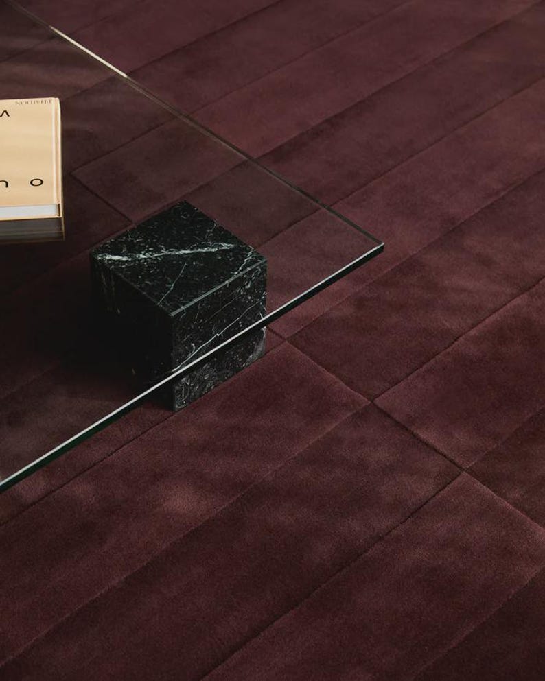 May include: A modern living room scene featuring a clear glass coffee table supported by a black marble cube. A book rests on the table, and the background is a burgundy, textured rug. The rug has a geometric pattern.