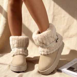May include: Beige winter boots with faux fur trim and a buckle detail. The boots have a platform sole and a soft, neutral color palette. The boots are designed for cold weather and casual wear.